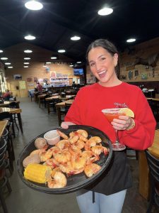 waitress in a red shirt holding a martini glass in one hand an a tray of boiled shrimp in the other