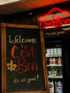 Welcome to C'est Bon chalkboard sign with neon sign in the background