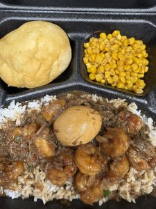 plate lunch featuring shrimp in a gravy over rice with a boiled egg, roll, and corn