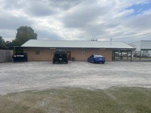 exterior image of tan brick building with a metal roof and gravel parking lot