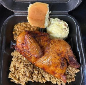 plate lunch featuring barbeque chicken, rice dressing, pork n beans, potato salad, and a roll