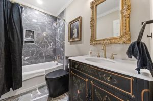 bathroom with ornately-painted vanity, black toilet, golden-framed mirror and tiled tub