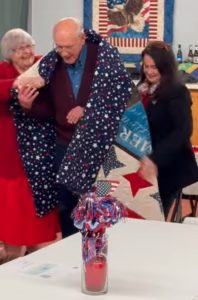 Two women on either side of an elderly gentlemen wrapping a quilt around his shoulders
