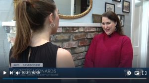Two women standing in front of a fireplace mantle for an interview, camera sees the back of one and face of the other