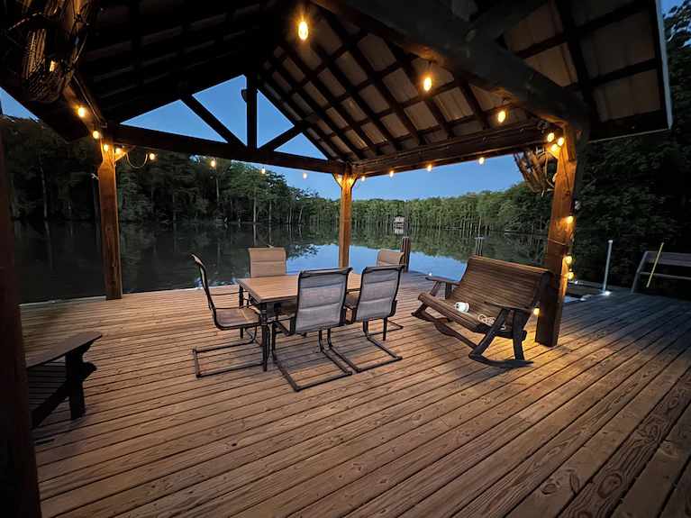 dusk photo of pavilion on the water with lights and an outdoor table and chairs