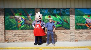 Artist Robert Dafford posing with Piggly Wiggly mascot in front of new frog murals in Rayne, Louisiana