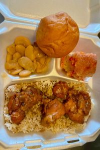 Plate lunch featuring meat, rice, sides, and a roll
