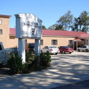brick church building with a red roof in the background with a white church sign in the parking lot of the foreground